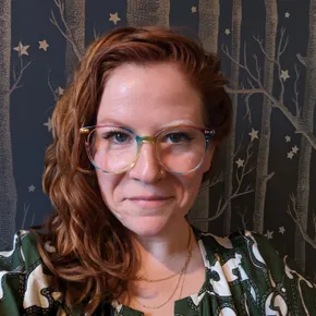 A photo of the artist - a redhead with rainbow glasses against a blue and gold background